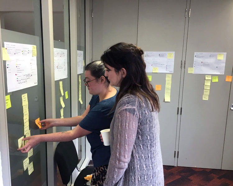 Two team members sorting and adding sticky notes to a glass wall of printed user journeys during a co-design workshop.