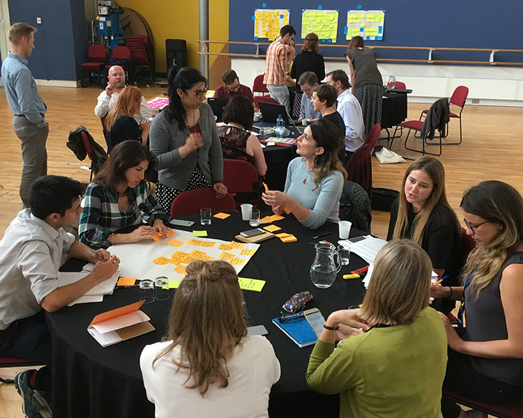 Large stakeholder workshop with multiple teams around tables, collaborating with sticky notes, worksheets, and group discussion.